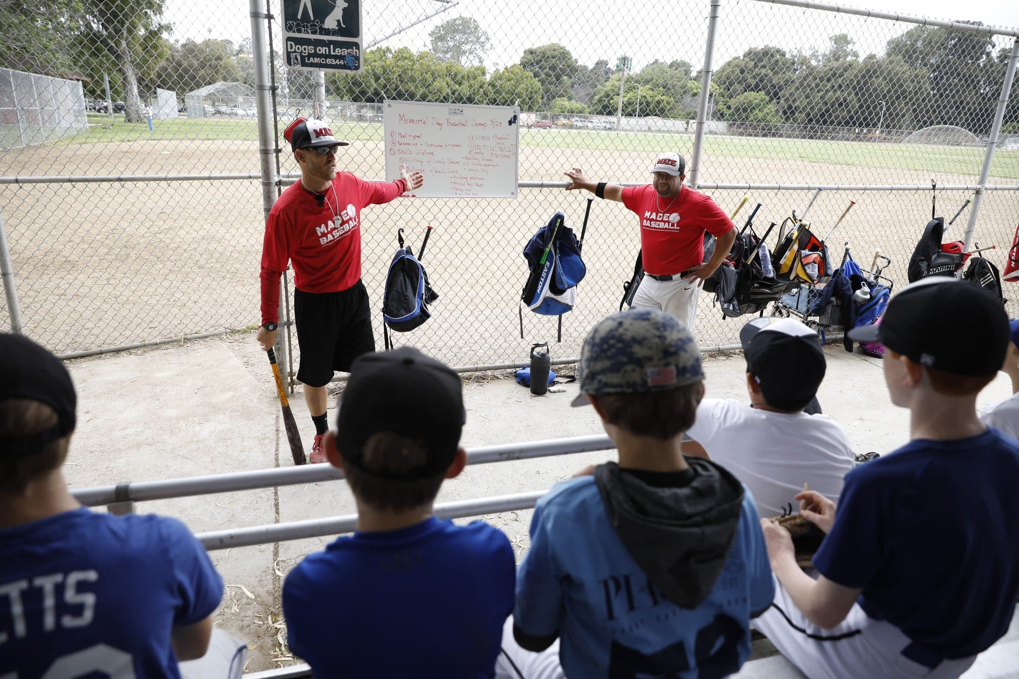 A MADE Baseball coach in a red shirt demonstrating to a row of seated campers, seen from behind, at a dugout