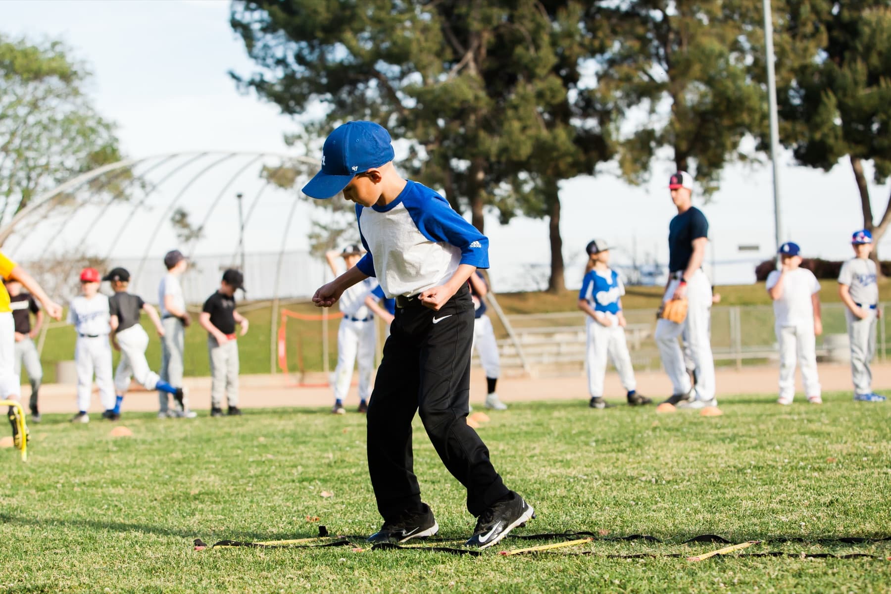A young camper in a blue cap works through a ladder footwork drill on the grass while other players wait their turn