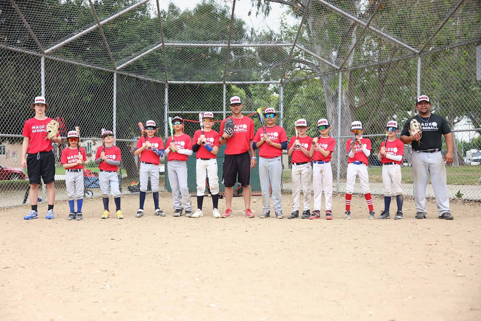 A line of campers in red MADE Baseball shirts standing along a fence with their coaches