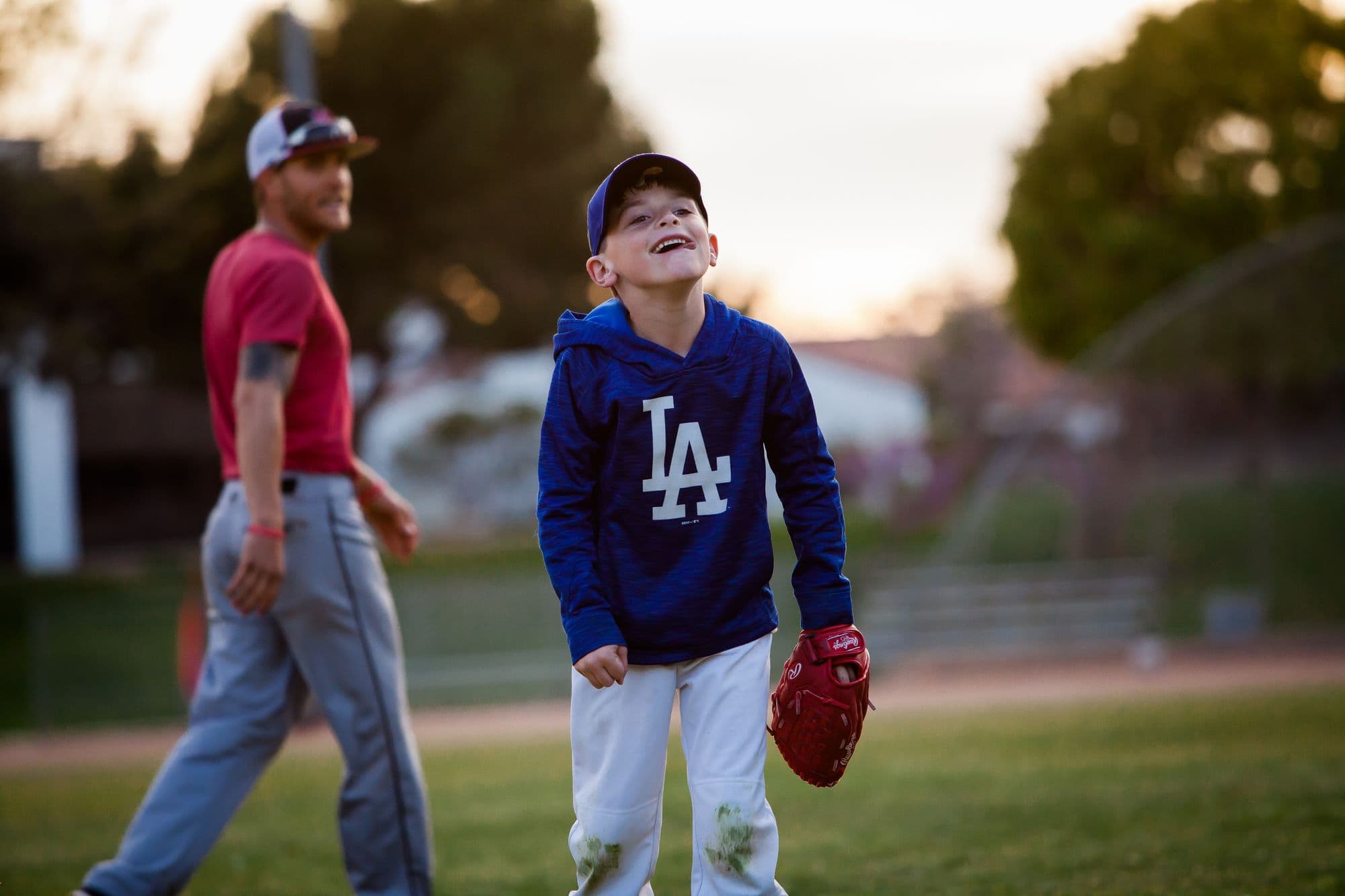 A young player grinning with a glove on during a camp session at dusk while a coach walks past