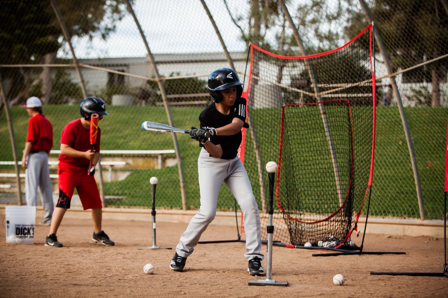 A young hitter swings off a batting tee in front of a net at a camp hitting station while another waits