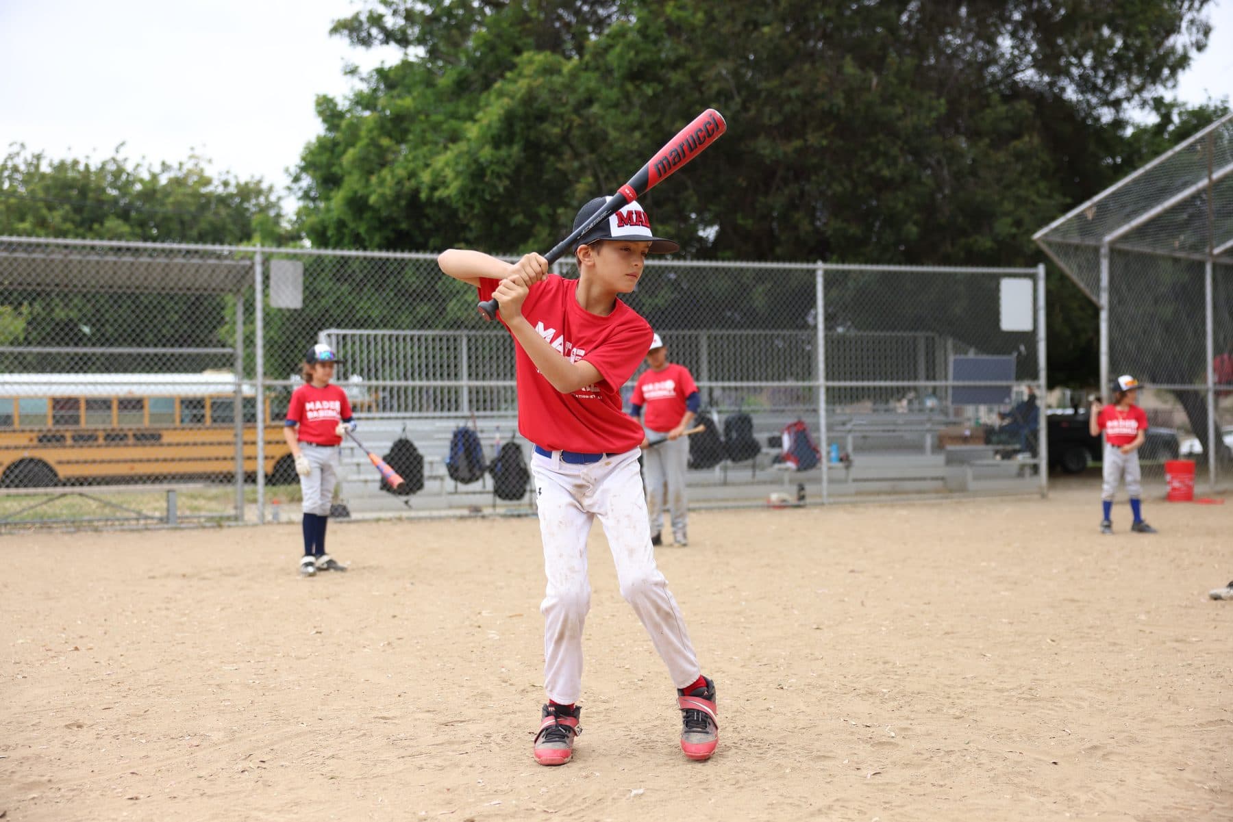 A young player in a red MADE Baseball shirt and cap sets up in his batting stance with the bat up while other players take swings behind him