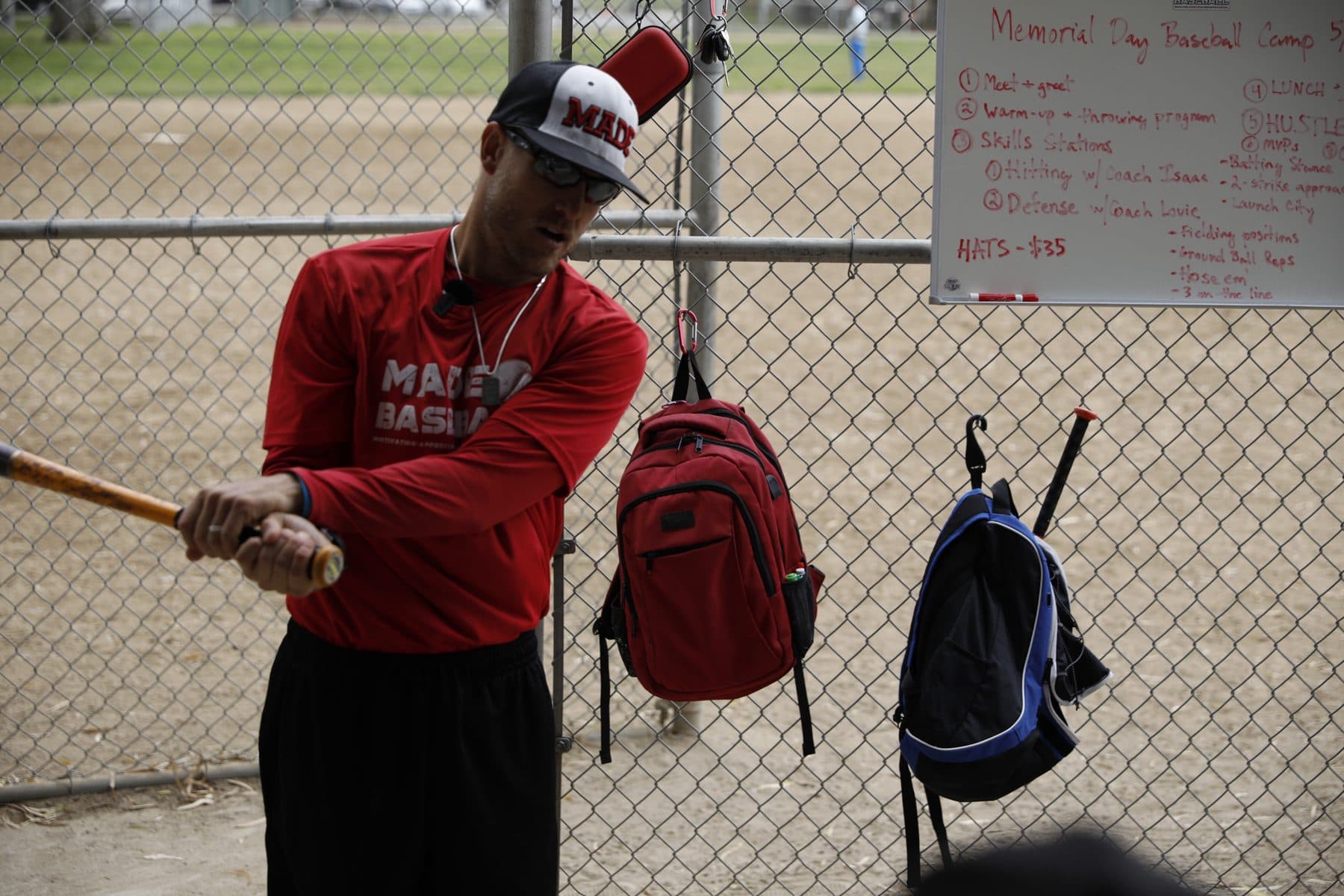 Coach Isaac Hess in a red MADE shirt and cap holding a bat in a close-up swing demonstration at a fence