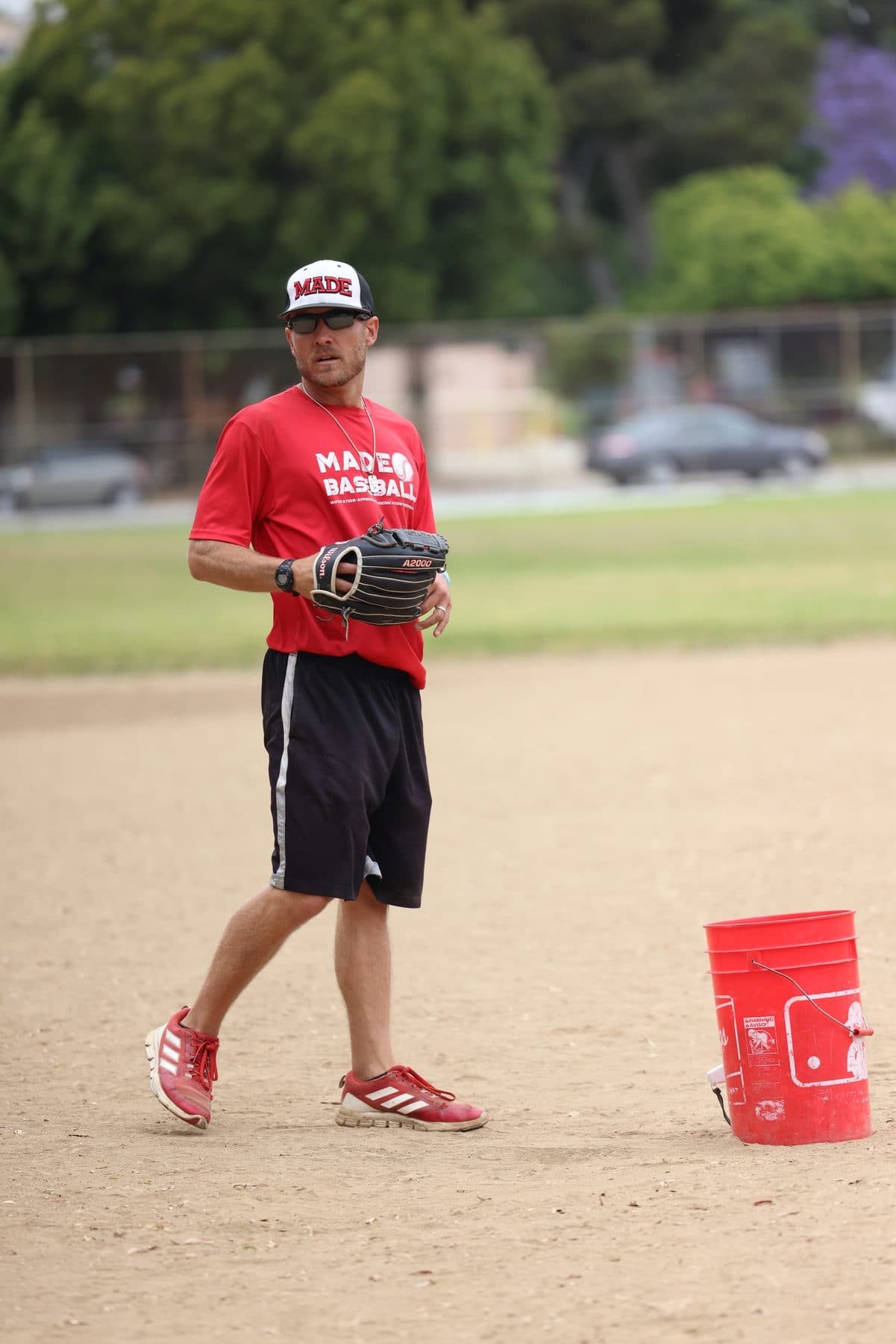 Coach Isaac Hess in a red MADE shirt, glove on, ready between drills with a ball bucket nearby