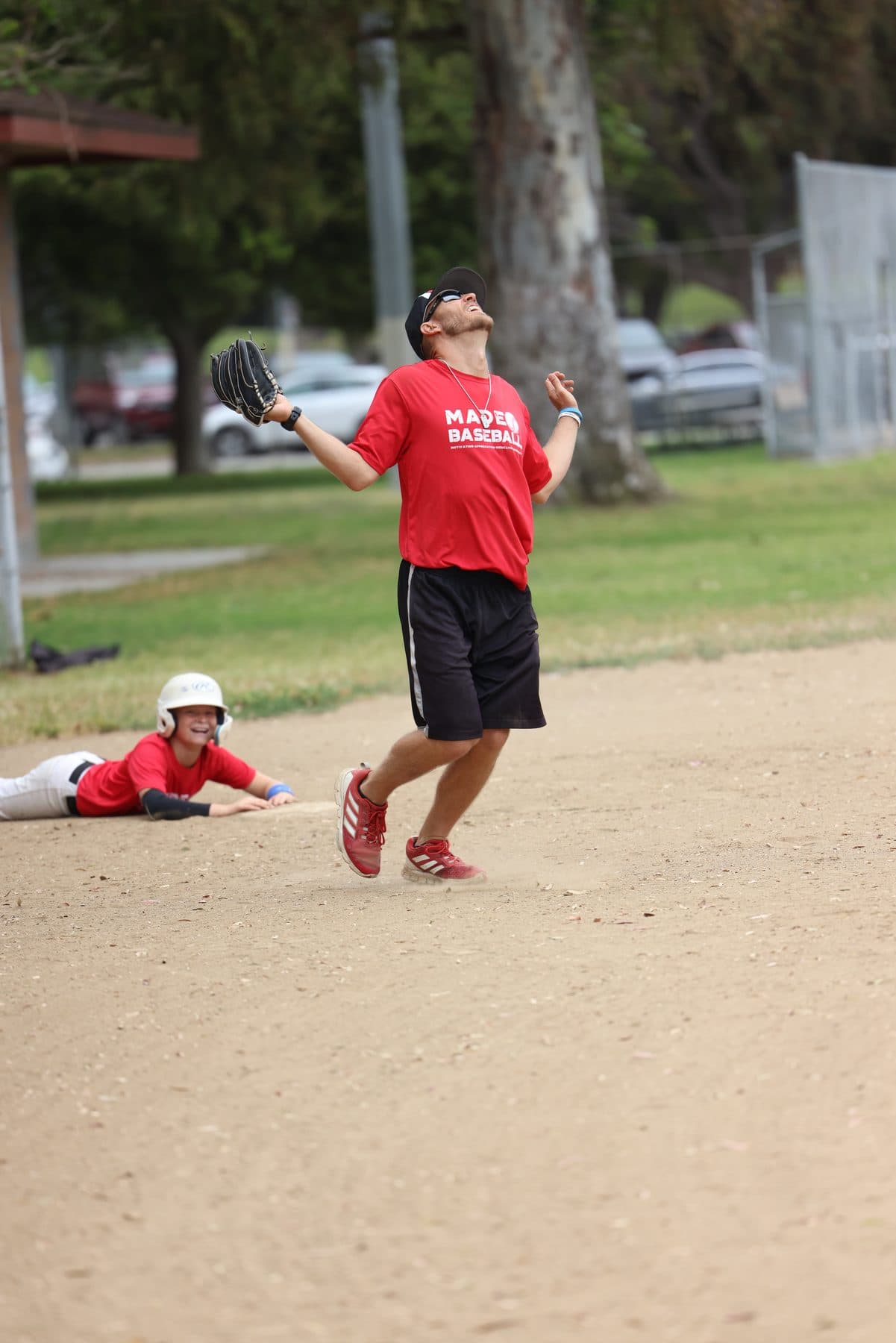 Coach Isaac Hess with his glove up looking skyward while a player slides into a base behind him