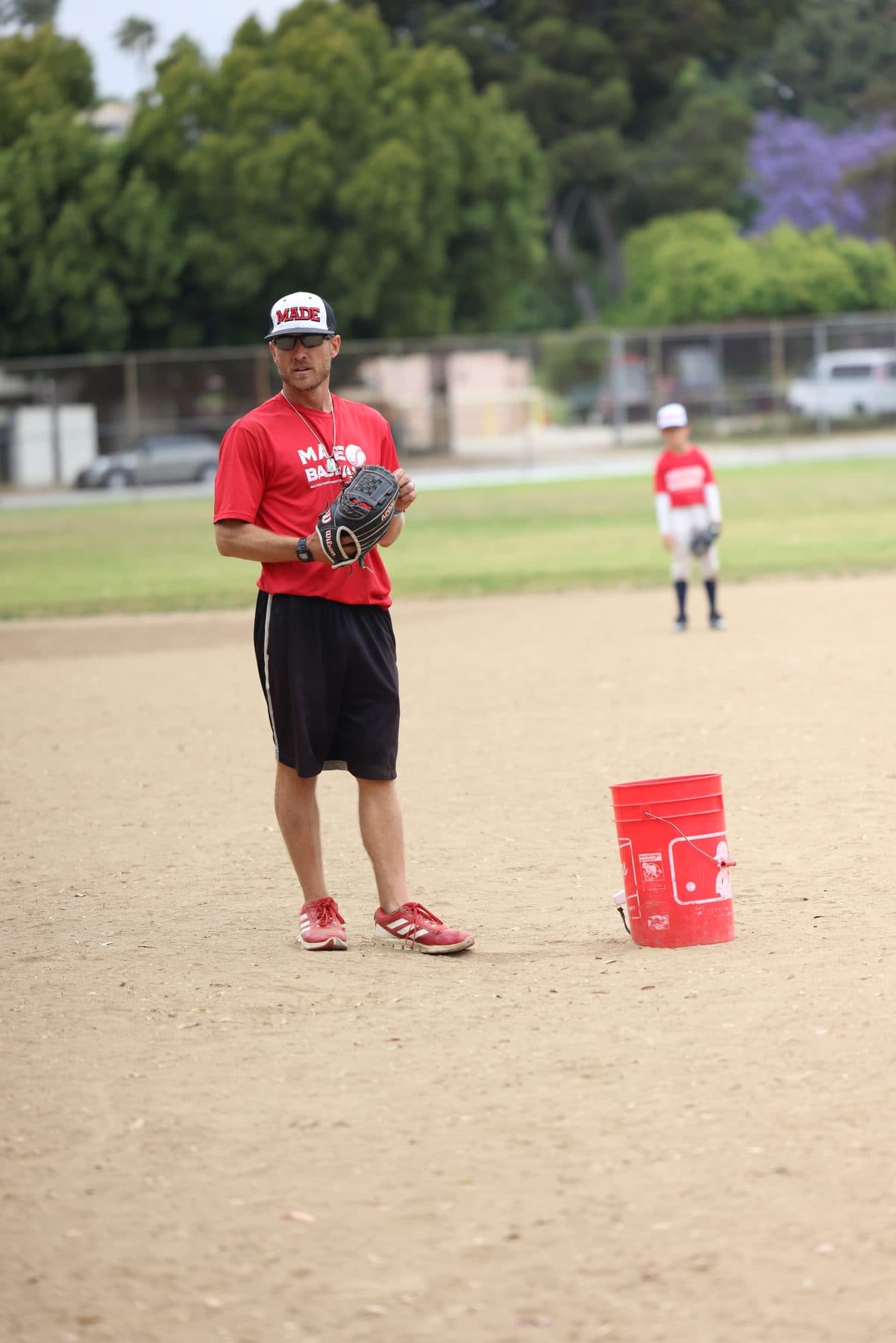 Coach Isaac Hess standing on a dirt infield with a glove on and a red bucket of balls beside him