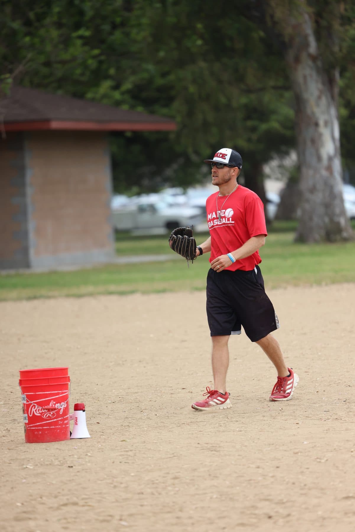 Coach Isaac Hess walking across a dirt infield with a glove, a red bucket and a megaphone behind him