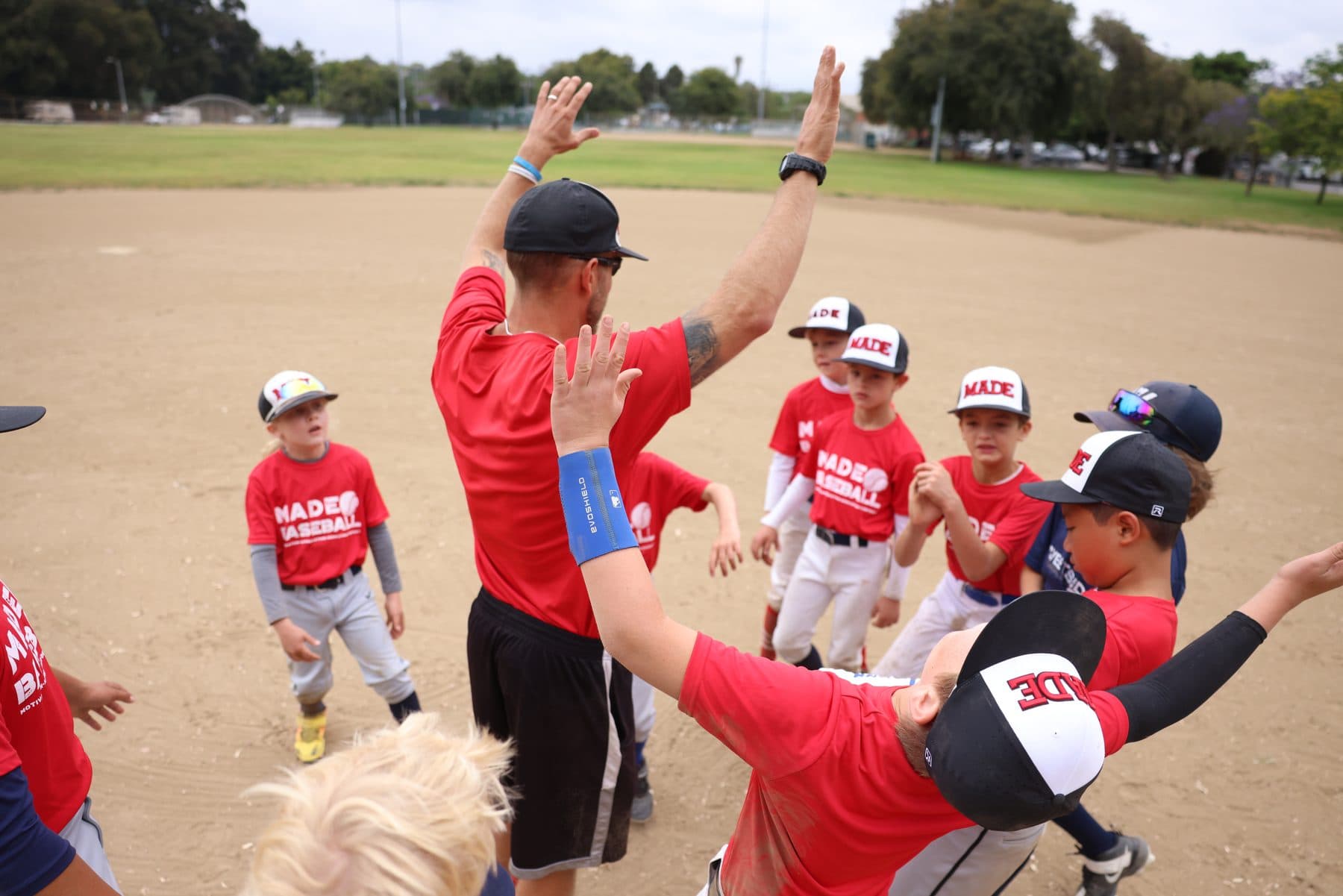Coach Isaac Hess in a red MADE Baseball shirt holds both hands high in the middle of a huddle of young players in red MADE shirts and caps