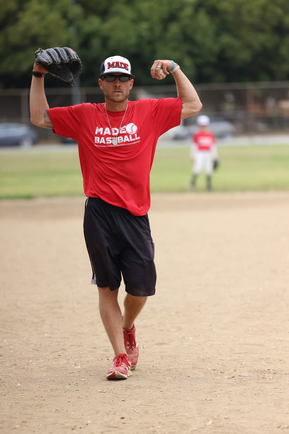 Coach Isaac Hess in a red MADE Baseball shirt and cap flexes both arms with a big grin on a baseball field