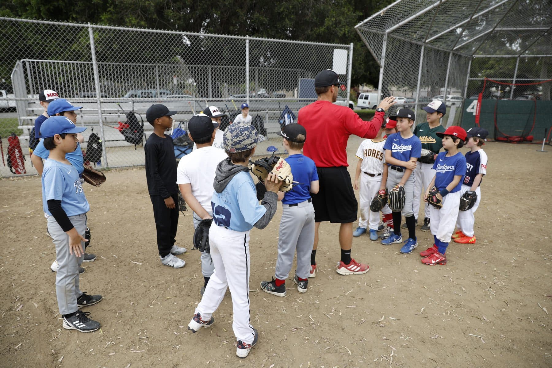 Coach Isaac Hess in a red shirt talks with a circle of young players holding gloves near a backstop