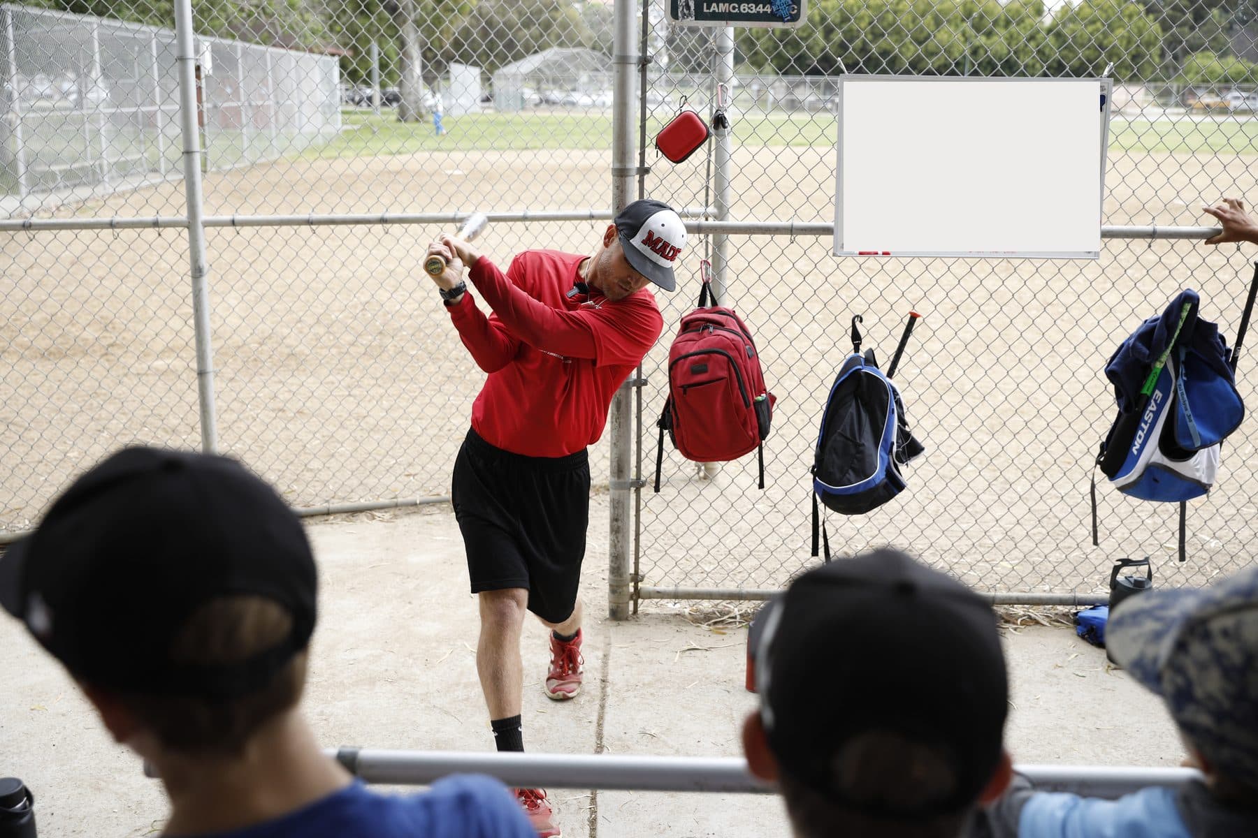 Coach Isaac Hess in red follows through on a demonstration swing while young players watch from the bench with their backs to the camera, backpacks hanging on the fence behind him