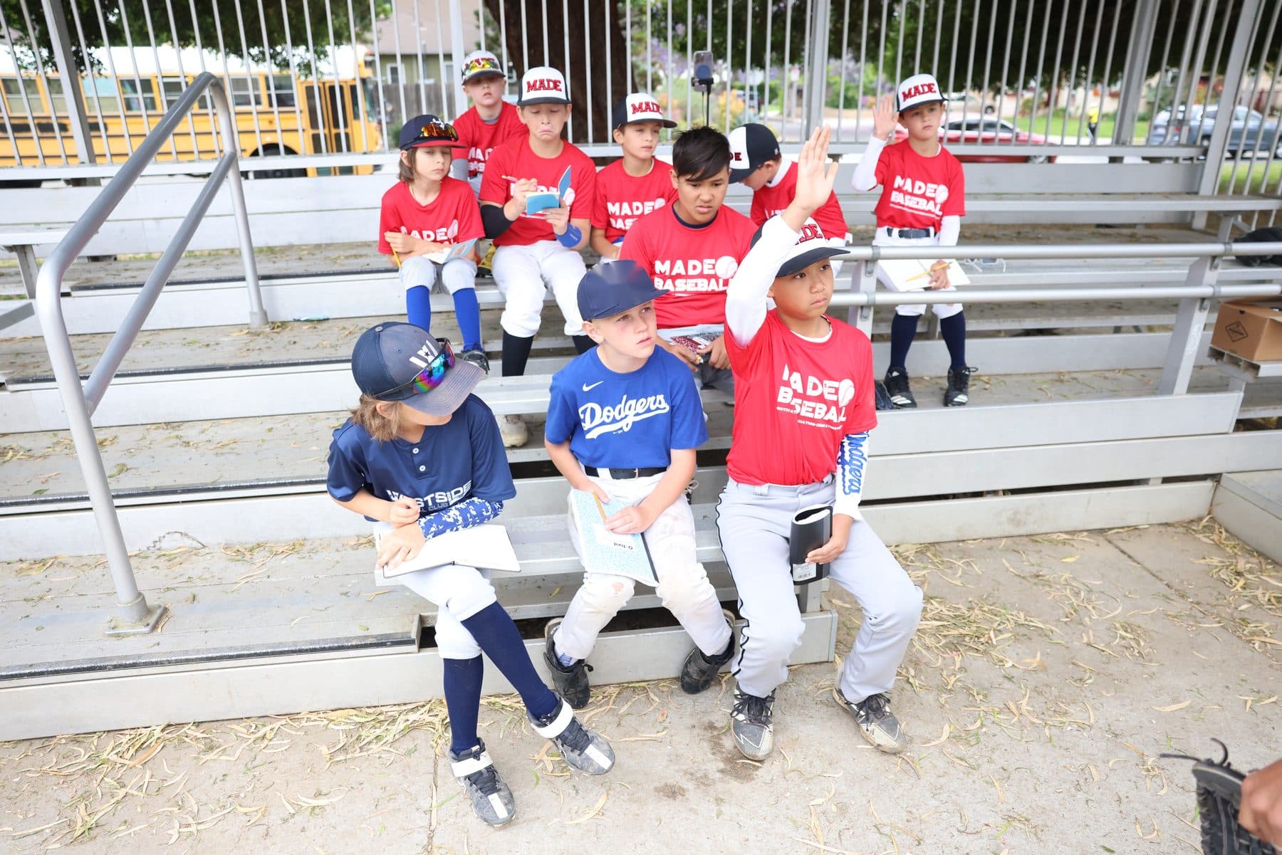 Young players in red MADE Baseball shirts and mixed team jerseys sit on the bleachers with notebooks and pencils, one with a hand raised