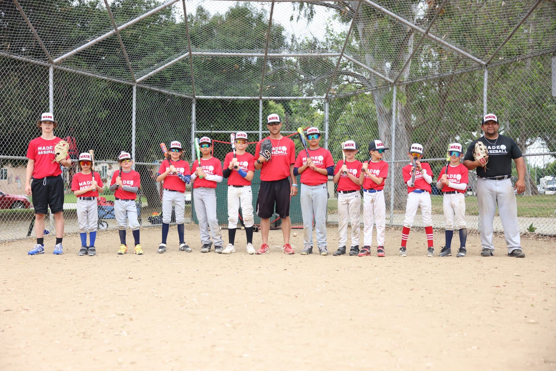 Coach Isaac Hess and a line of young players in red MADE Baseball shirts hold their bats upright on the infield dirt in front of a backstop