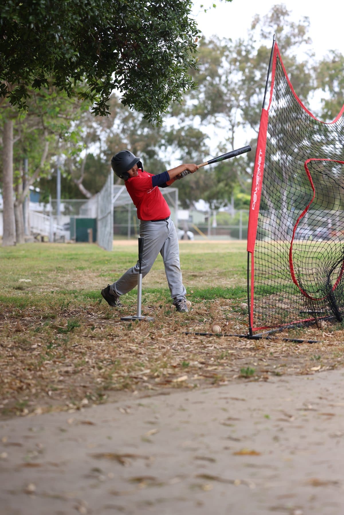 A young hitter mid-swing off a tee beside a red practice net under trees