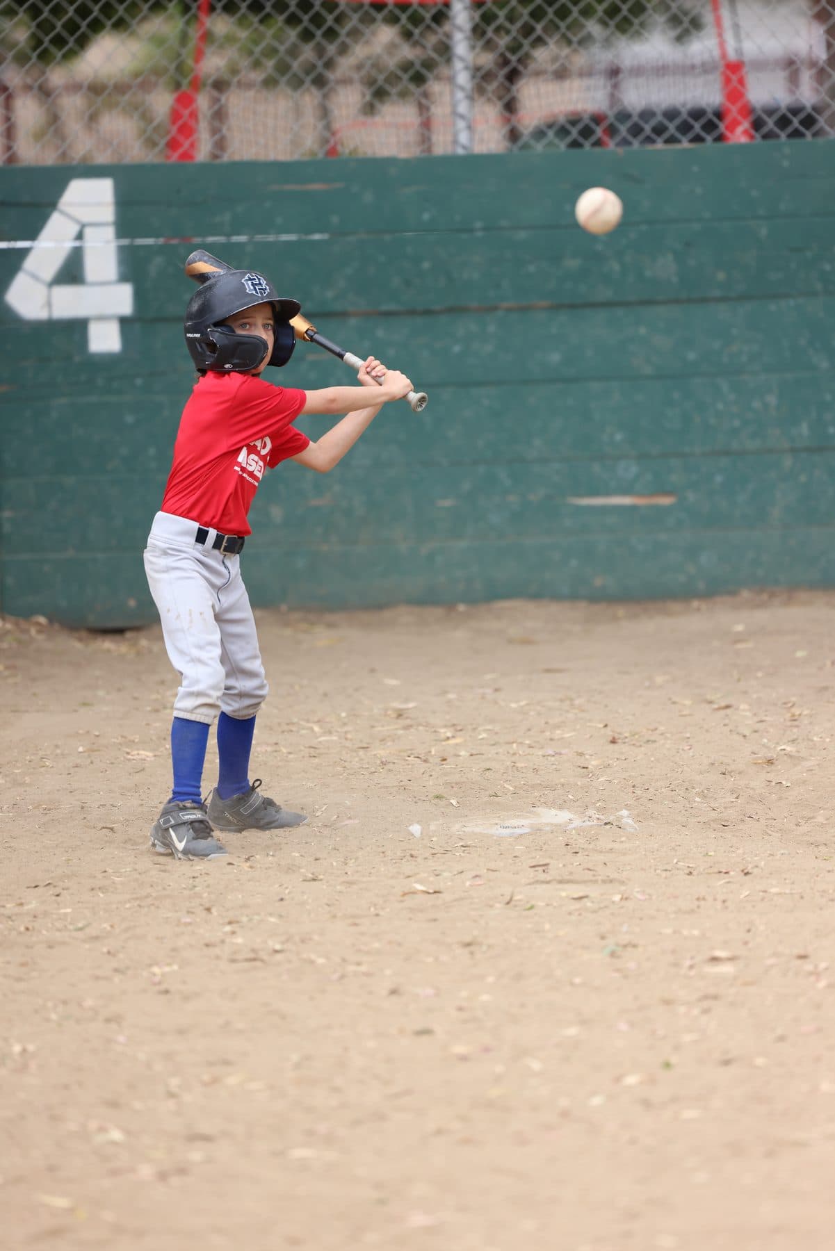 A young hitter in a helmet in his stance, eyes on the ball coming in, in front of a green backstop wall