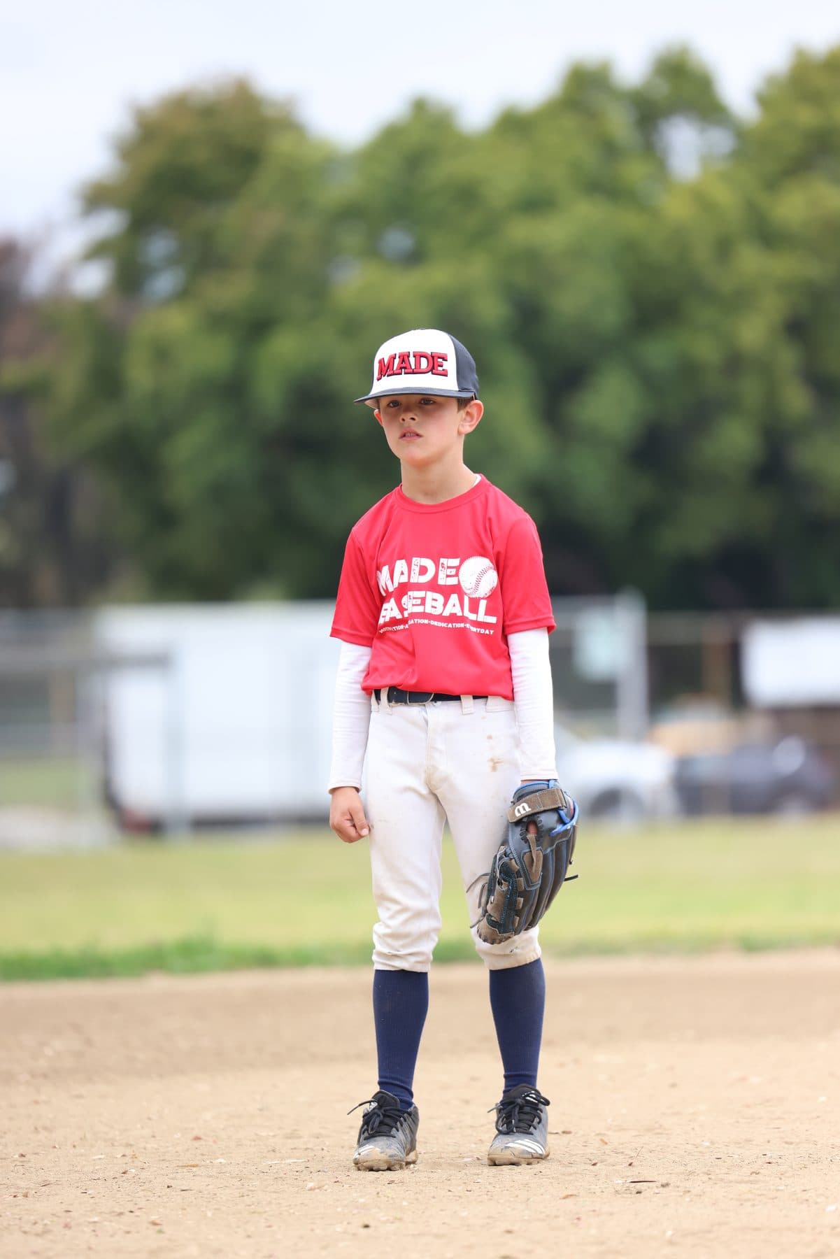A young player in a red MADE shirt and cap standing ready with his glove at his side
