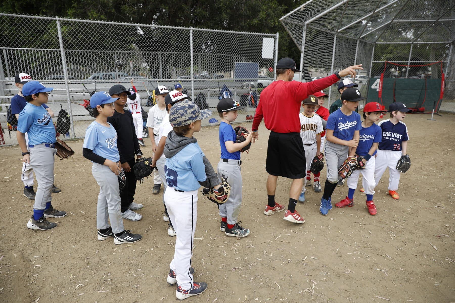 Coach Isaac Hess in a red shirt points across the field while a big group of young players in gloves and mixed team jerseys gathers around him near a backstop