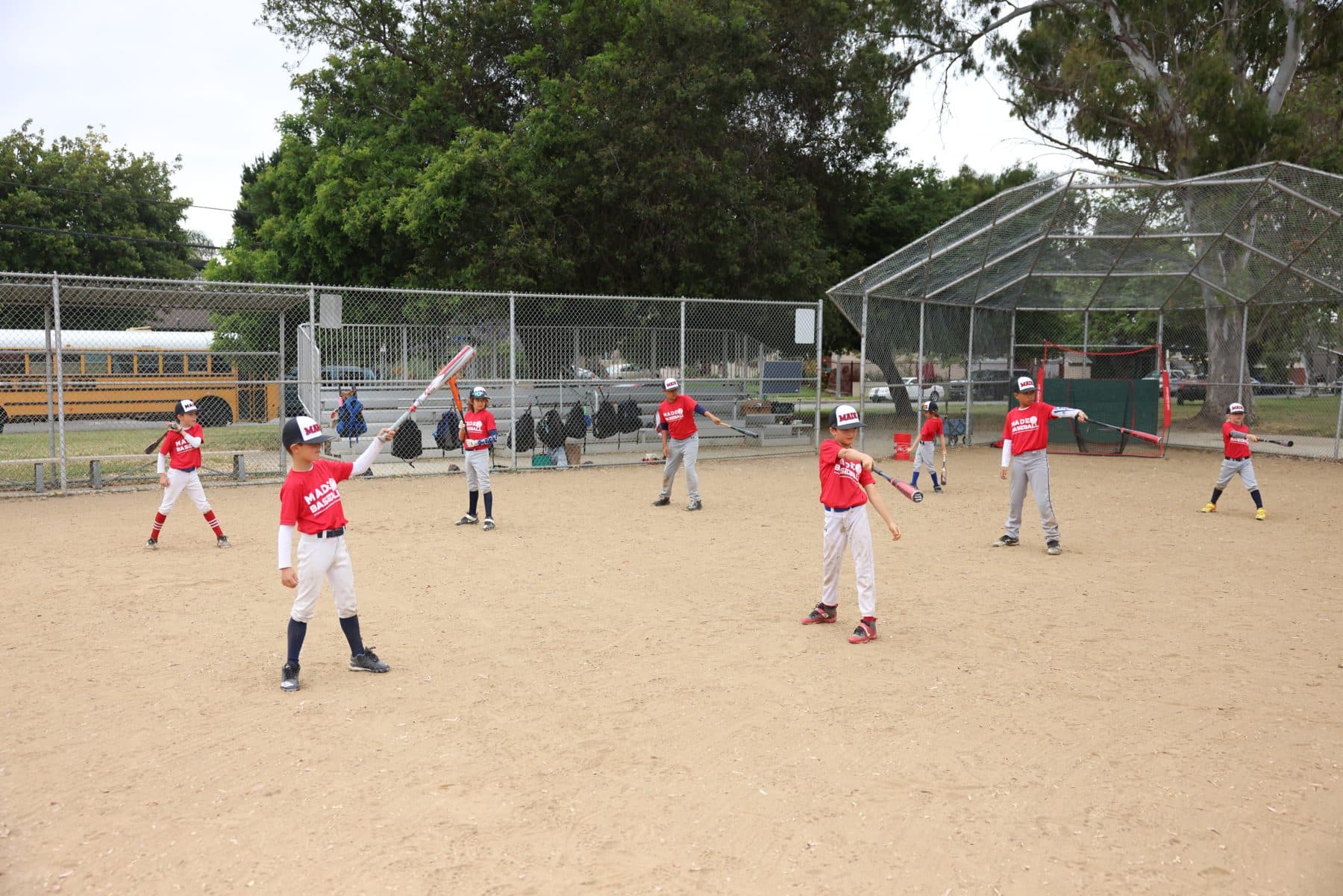 A group of young players in red MADE Baseball shirts spread across the infield dirt, all taking easy swings with their bats