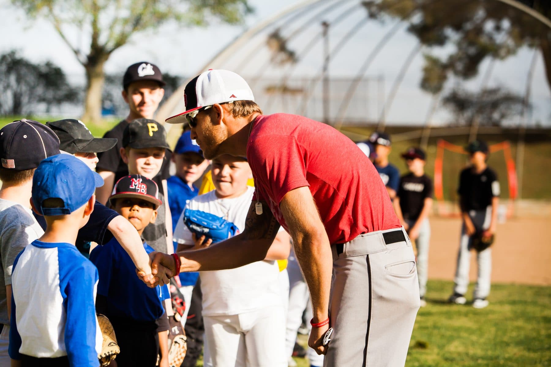 Coach Isaac Hess in a red shirt leans down to shake hands with a young player while a group of smiling kids in gloves and caps looks on