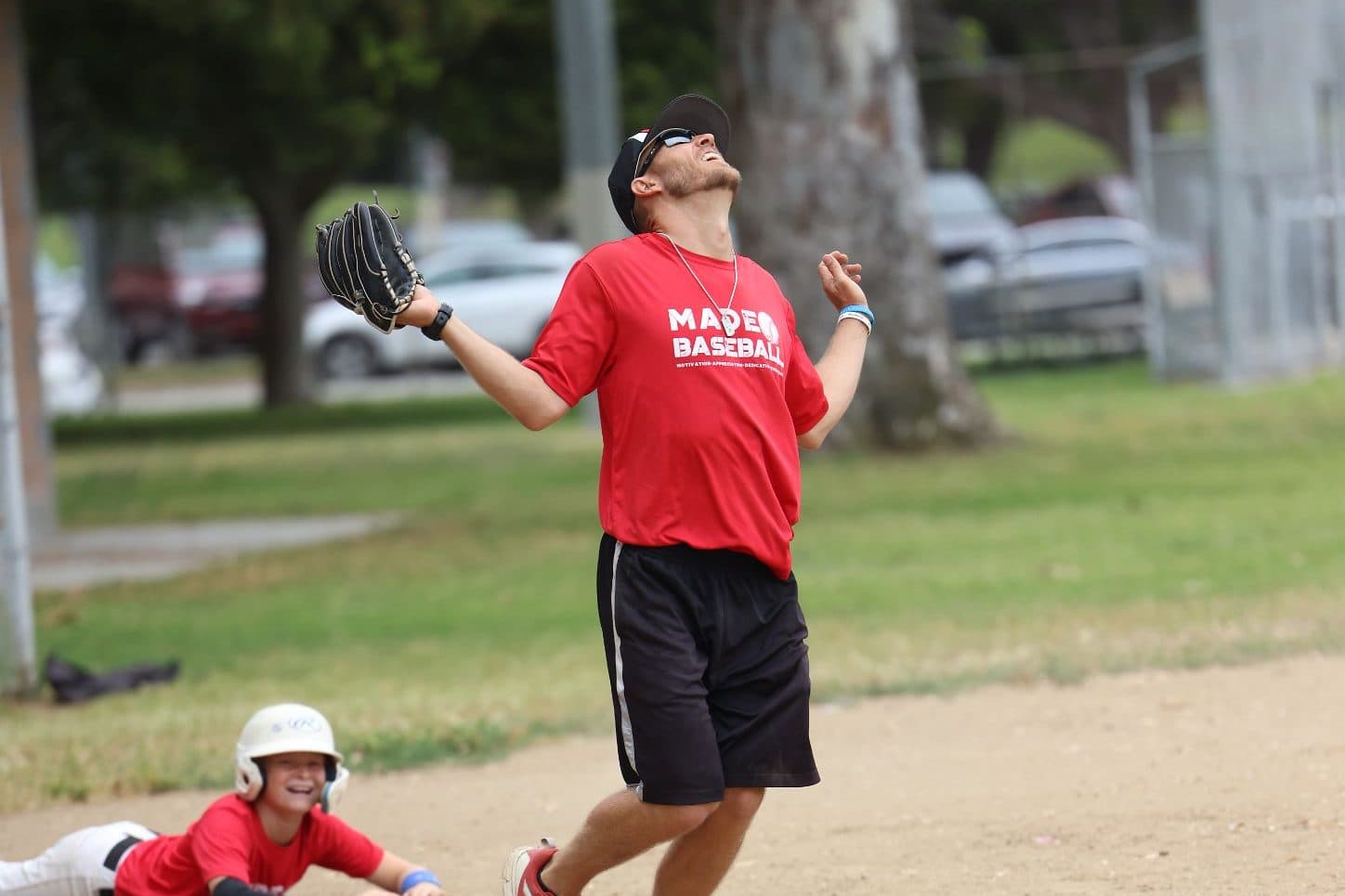 Coach Isaac Hess in a red MADE shirt looks up with his glove out for a pop fly while a young player in a helmet lies on the dirt behind him laughing
