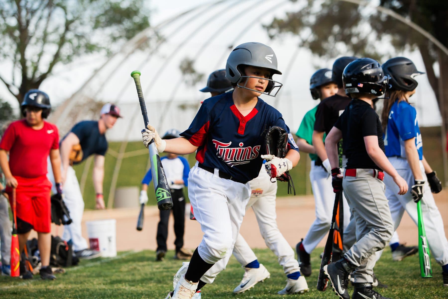A young player in a batting helmet jogs forward carrying a bat and glove while other kids walk past on the grass behind him