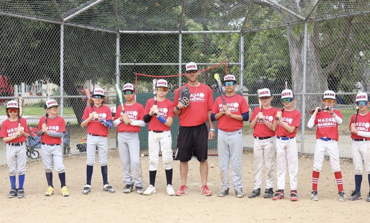 A MADE Baseball coach and a lineup of young players in matching red jerseys and caps, smiling on the field in front of a backstop