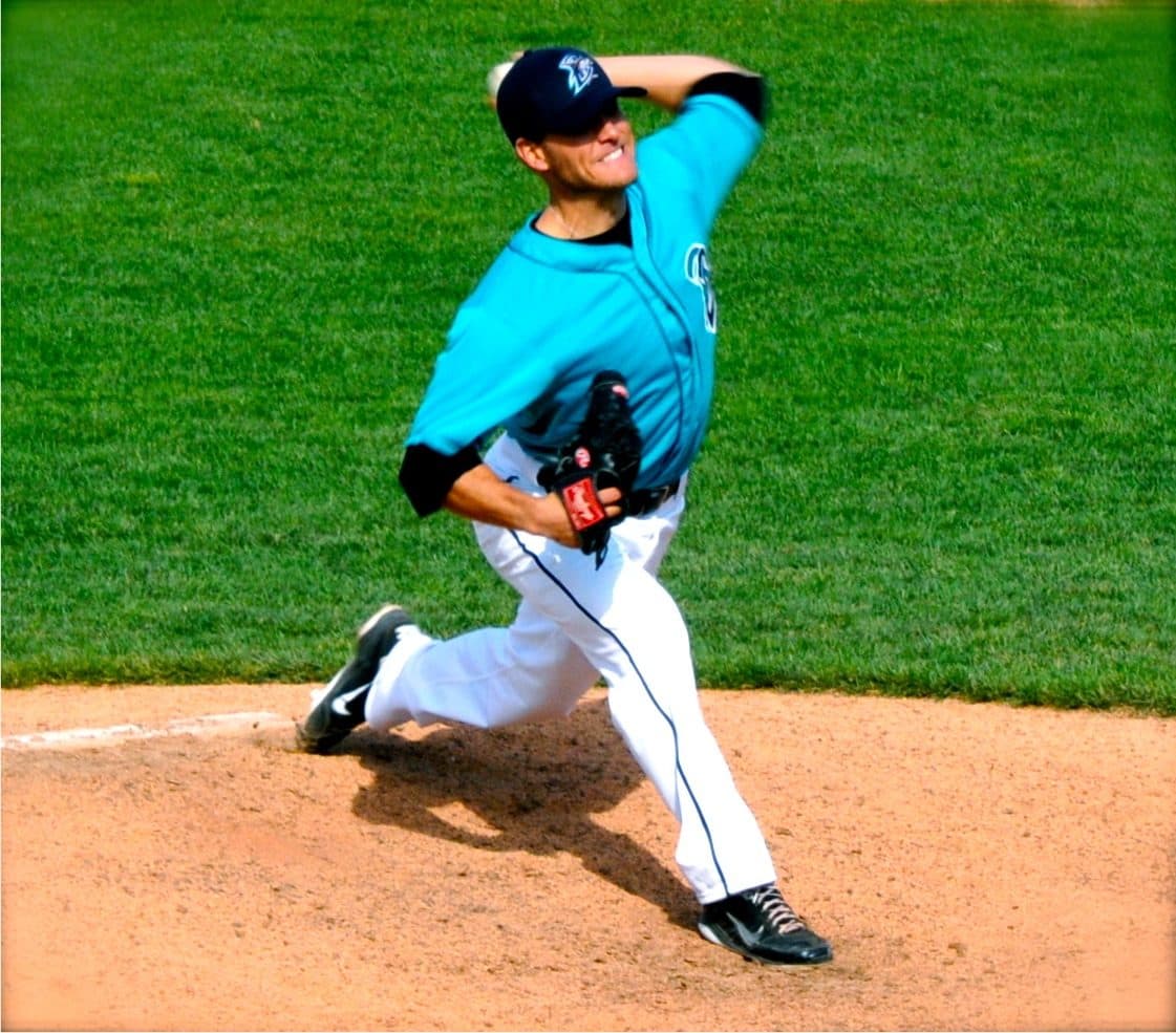 Coach Isaac Hess pitching left-handed from the mound in a teal jersey, mid-delivery