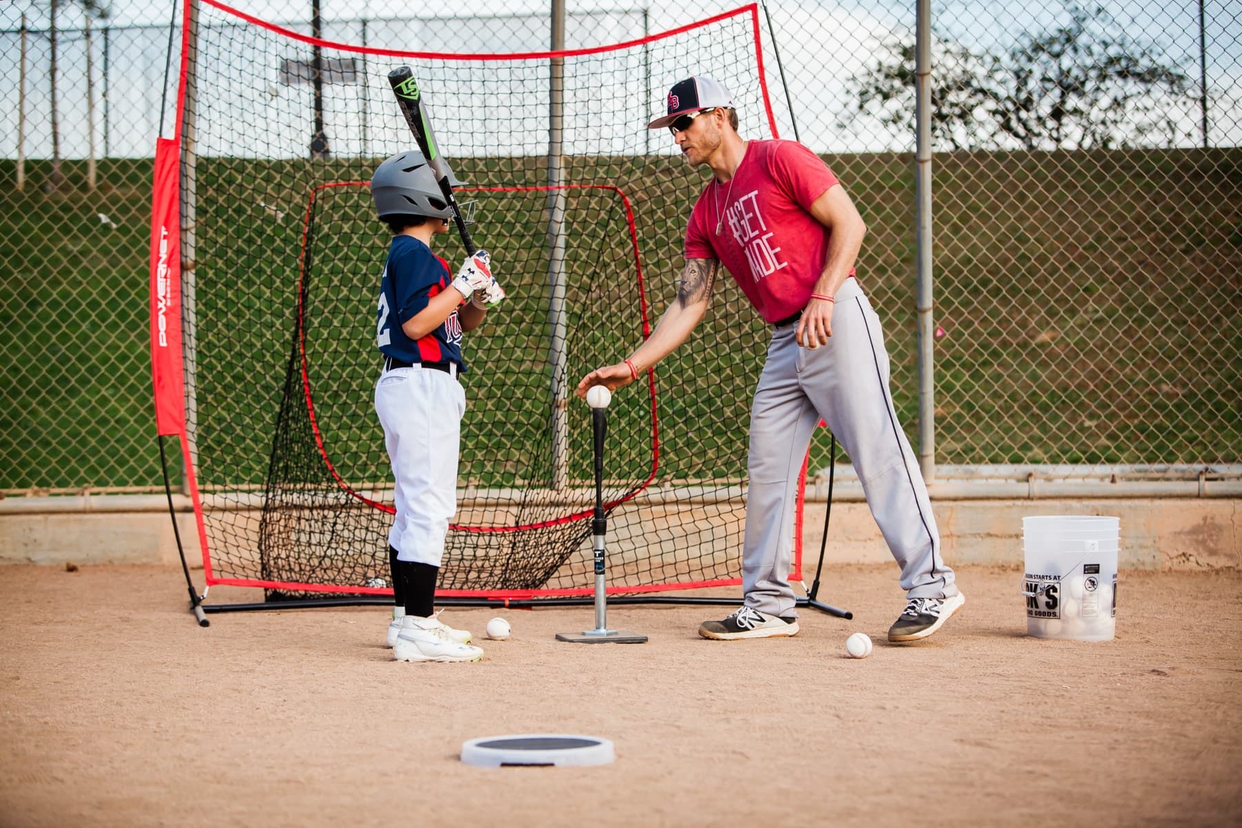 Coach Isaac Hess in a red #GETMADE shirt sets a baseball on a batting tee while a young hitter in a helmet waits in his stance in front of a net