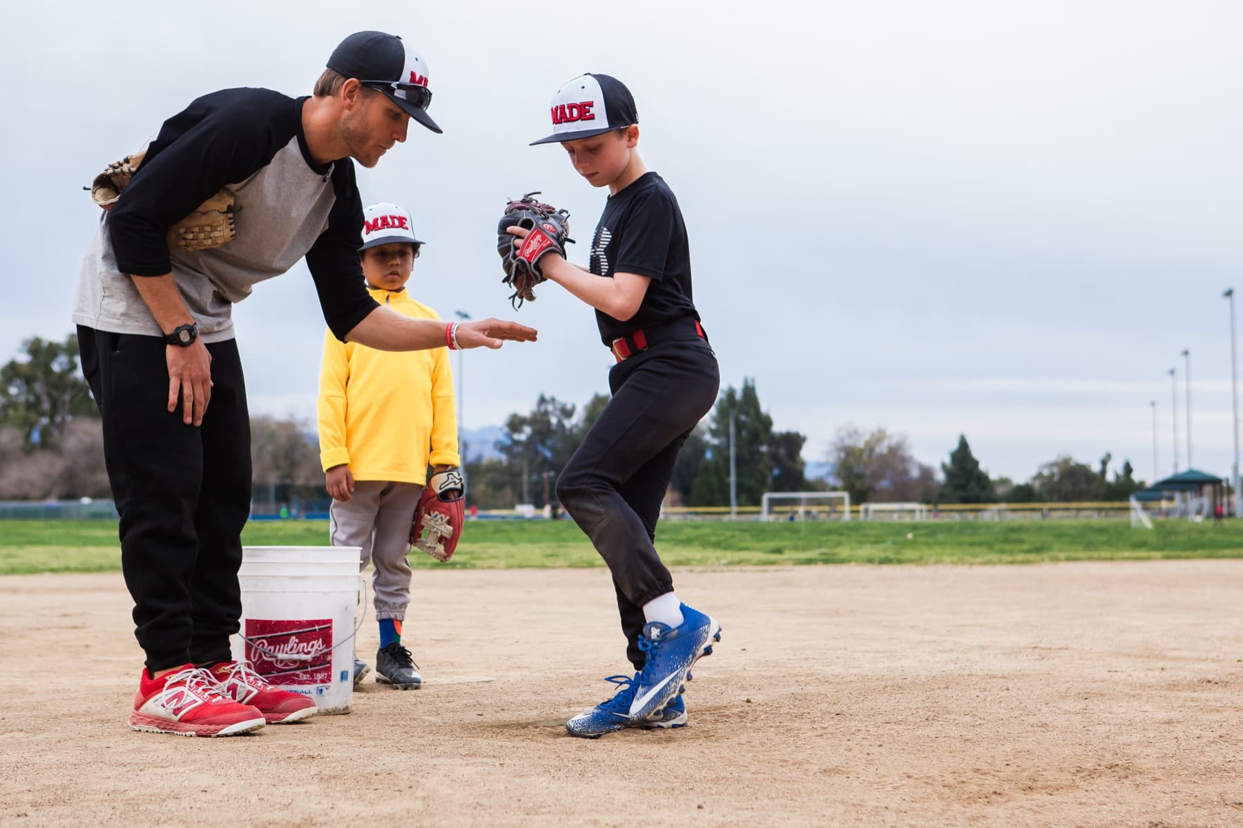 Coach Isaac Hess bending beside a young pitcher at the top of his leg lift while another player watches
