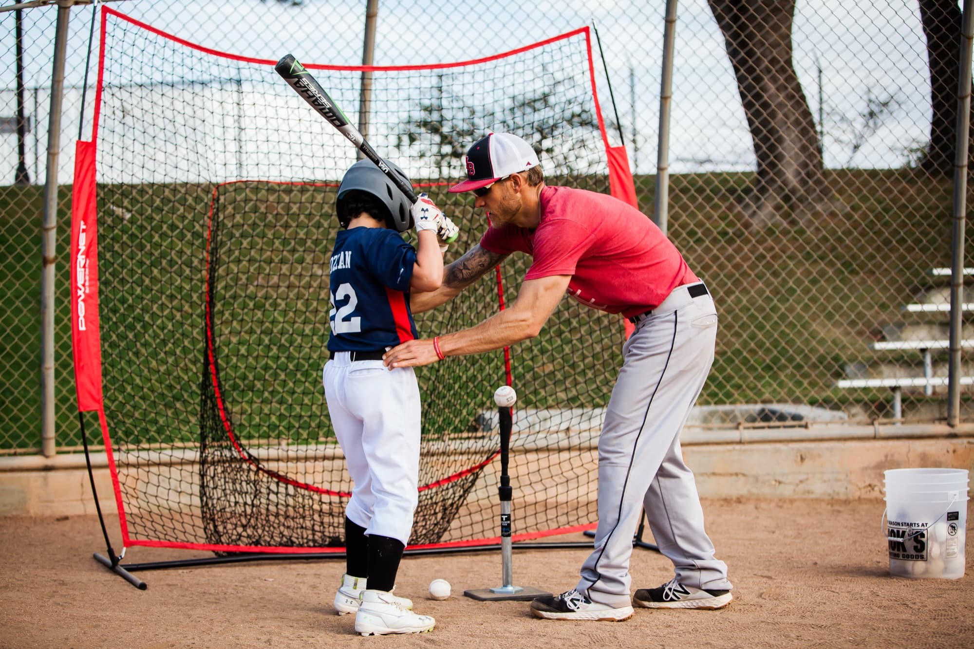 Coach Isaac Hess in a red shirt coaching a young hitter at a batting net, hands on the player's shoulders and bat