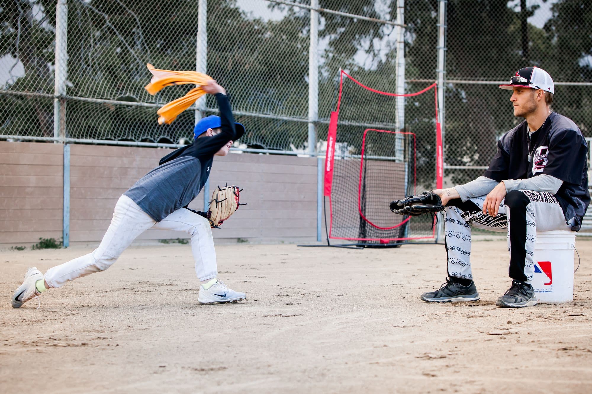 A young pitcher stretching into a delivery during a drill while Coach Isaac Hess watches from a bucket seat