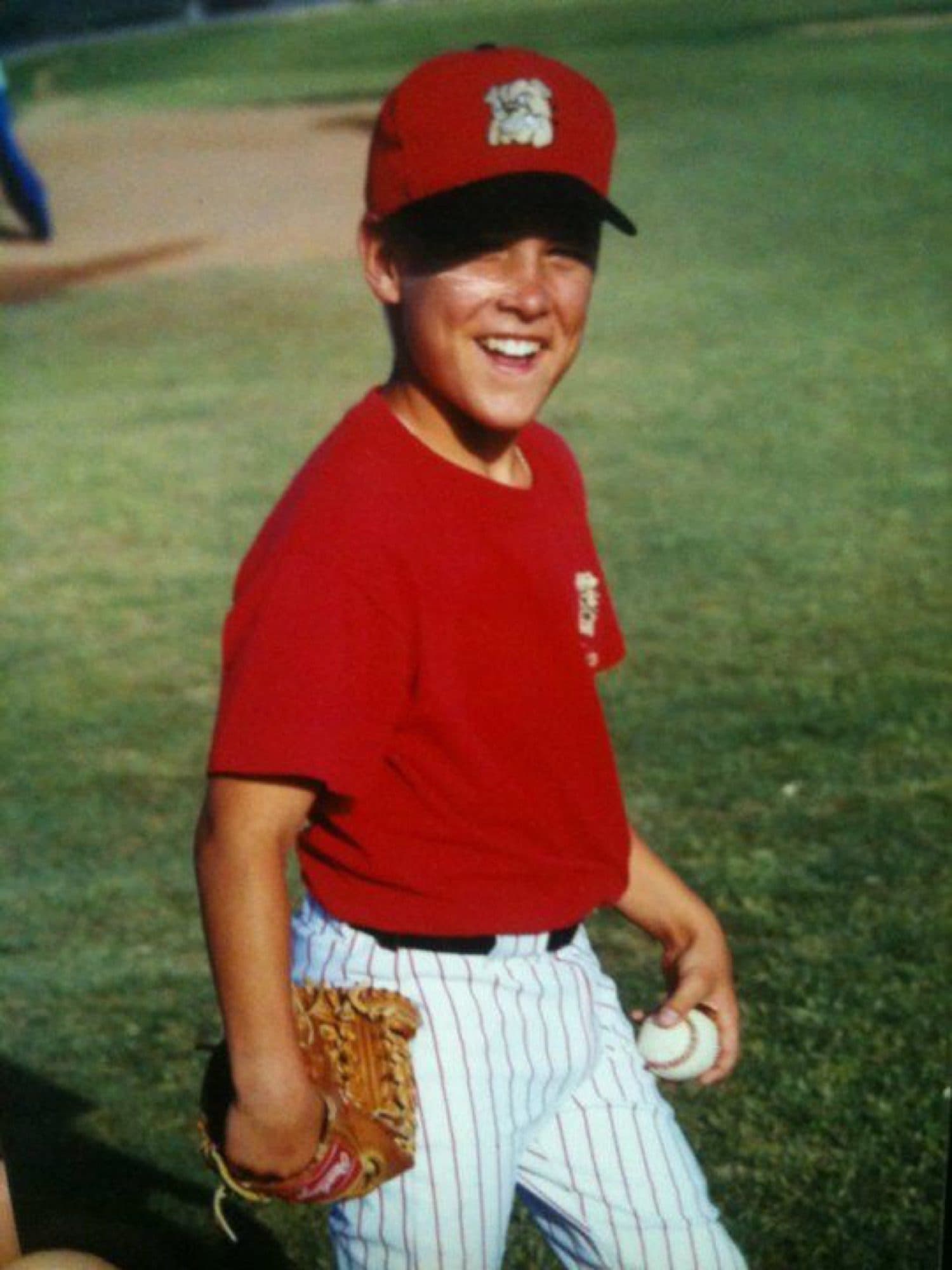 Coach Isaac Hess as a smiling kid in a red cap, red shirt and pinstripe pants, holding a glove and a baseball
