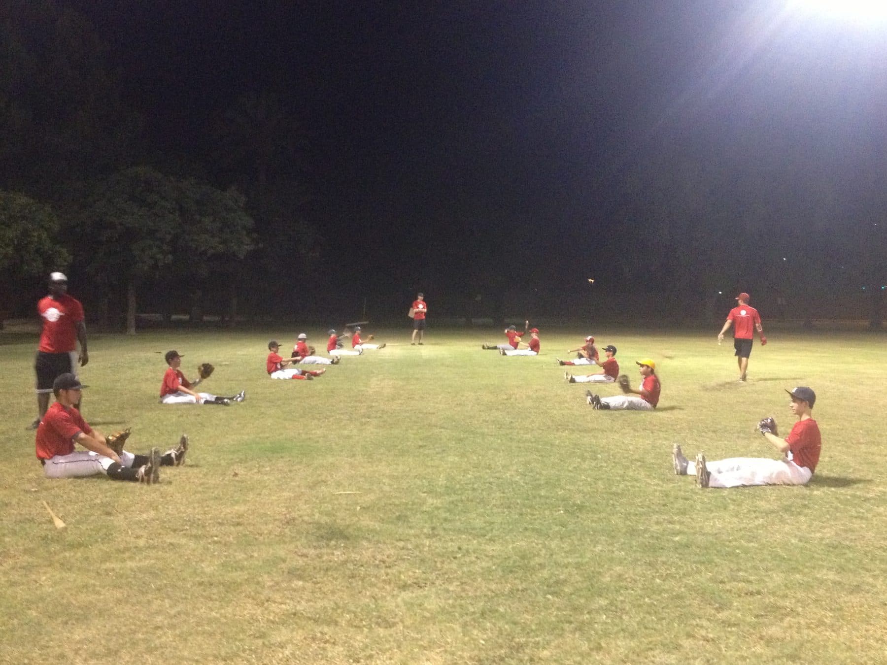 Players seated in a wide spread on the grass at night with gloves up, working a catching drill under field lights, a coach standing nearby