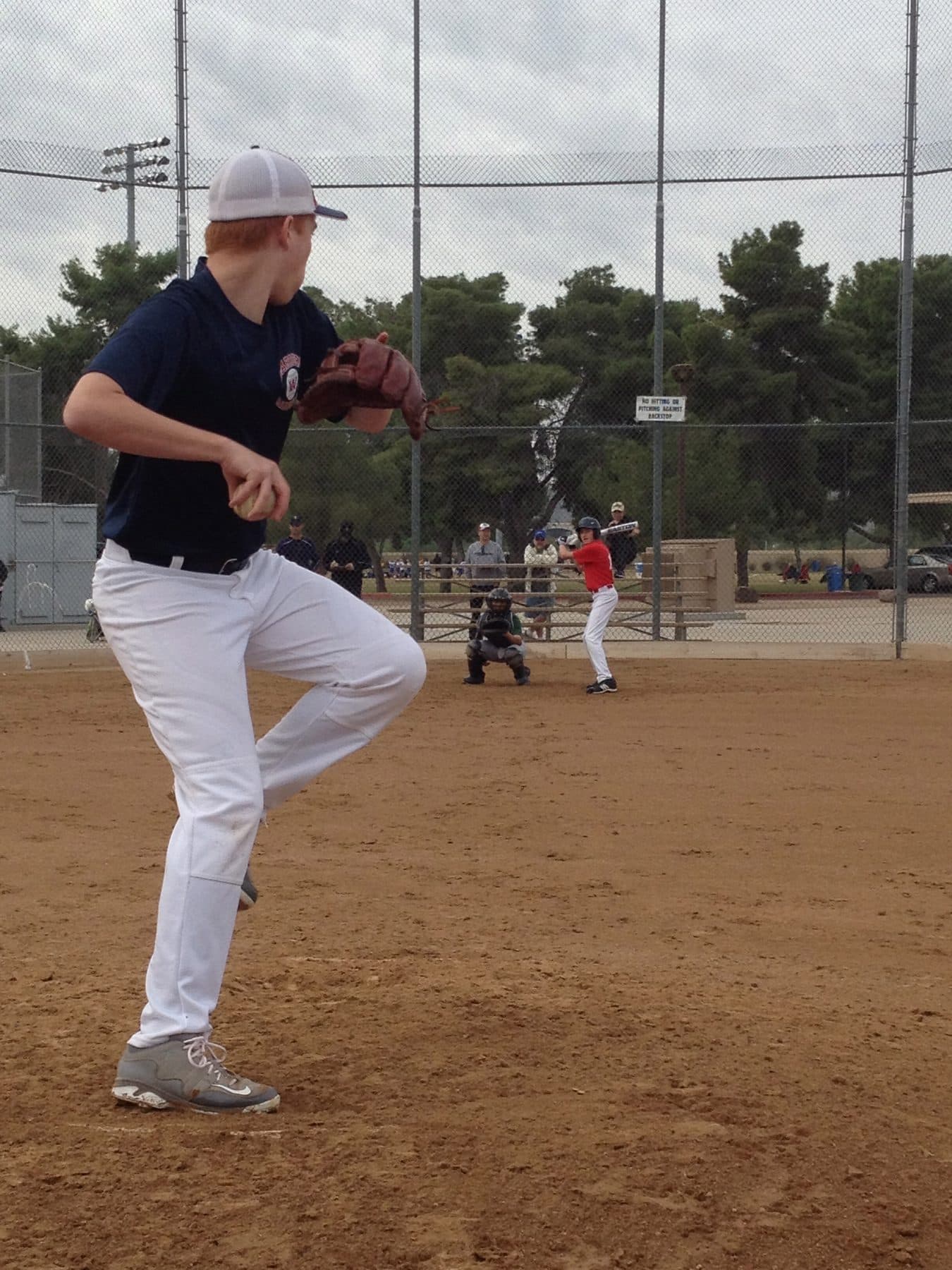 A teen pitcher at the top of his leg lift on a dirt mound, glove up, during practice