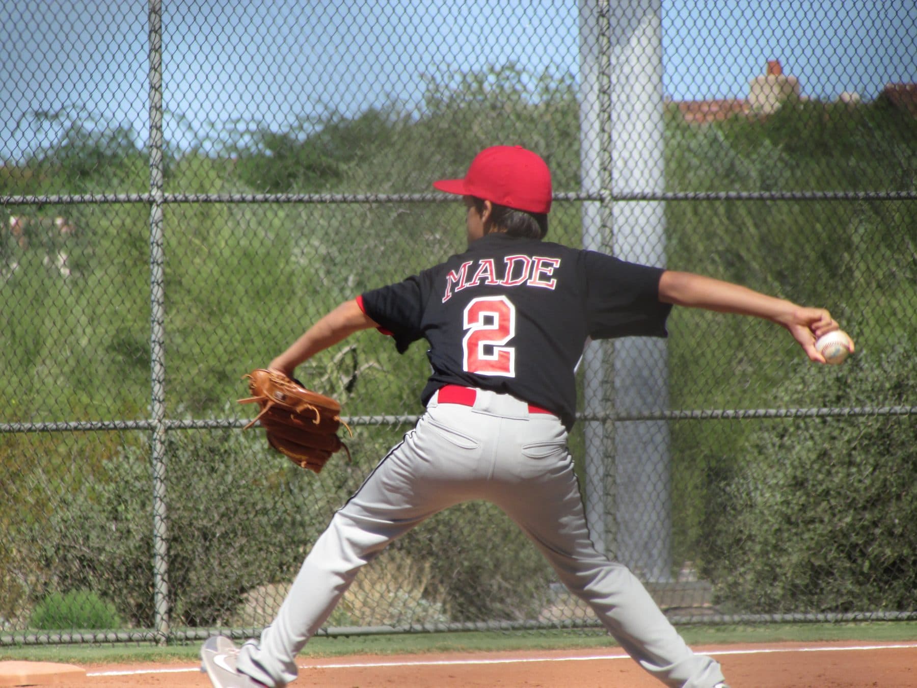 A young pitcher in a black MADE jersey, seen from behind, releasing the ball from the mound